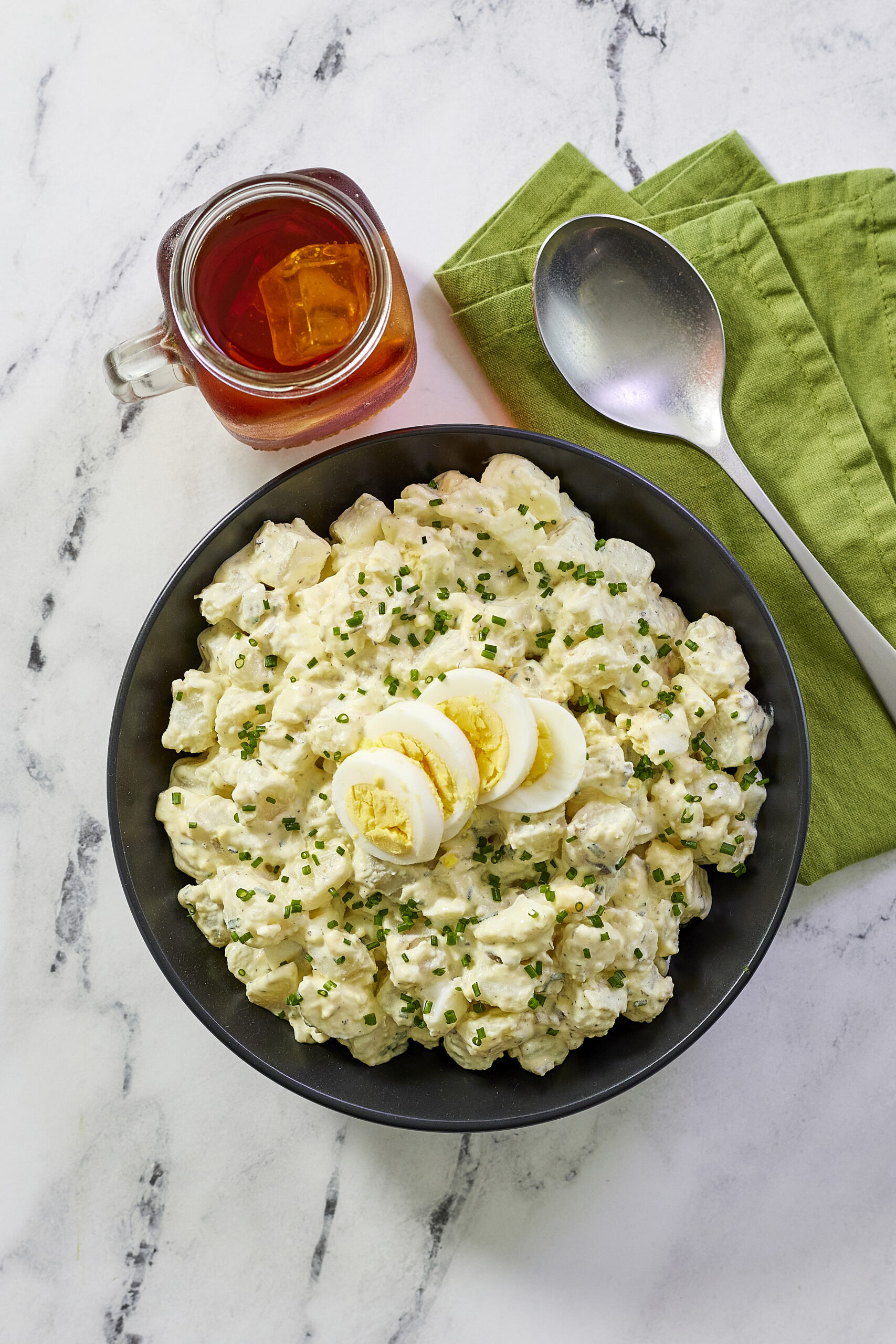 overhead shot of a bowl of potato salad with a glass of sweet iced tea and a silver spoon nearby.